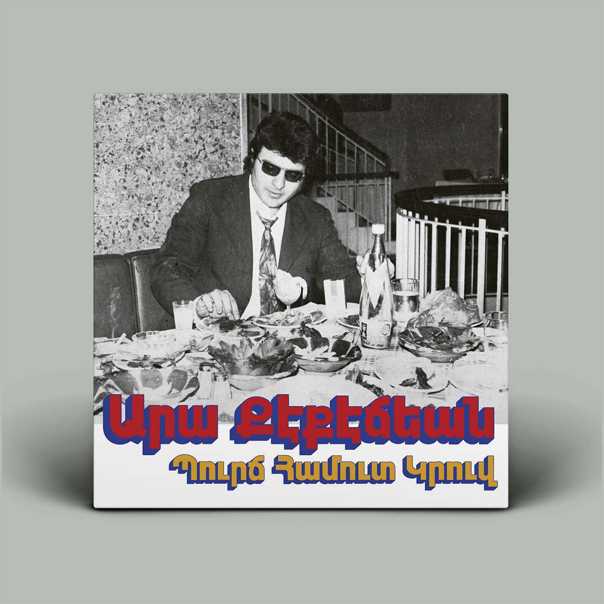 Album cover with a black and white photo of a man at a table with food, featuring colorful text at the bottom.