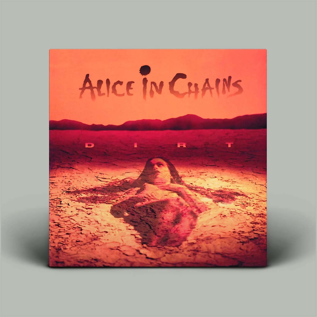 A square album cover for 'Dirt' by the band 'Alice in Chains', displayed against a neutral gray background with a soft drop shadow. The artwork features a surreal desert landscape under a glowing orange sky, with a woman's face emerging from cracked earth.