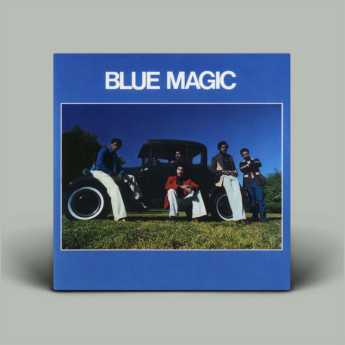 A square album cover for 'Blue Magic' featuring a blue border with white text and a central photograph of five people posing with an old car on grass, shown against a light gray background.