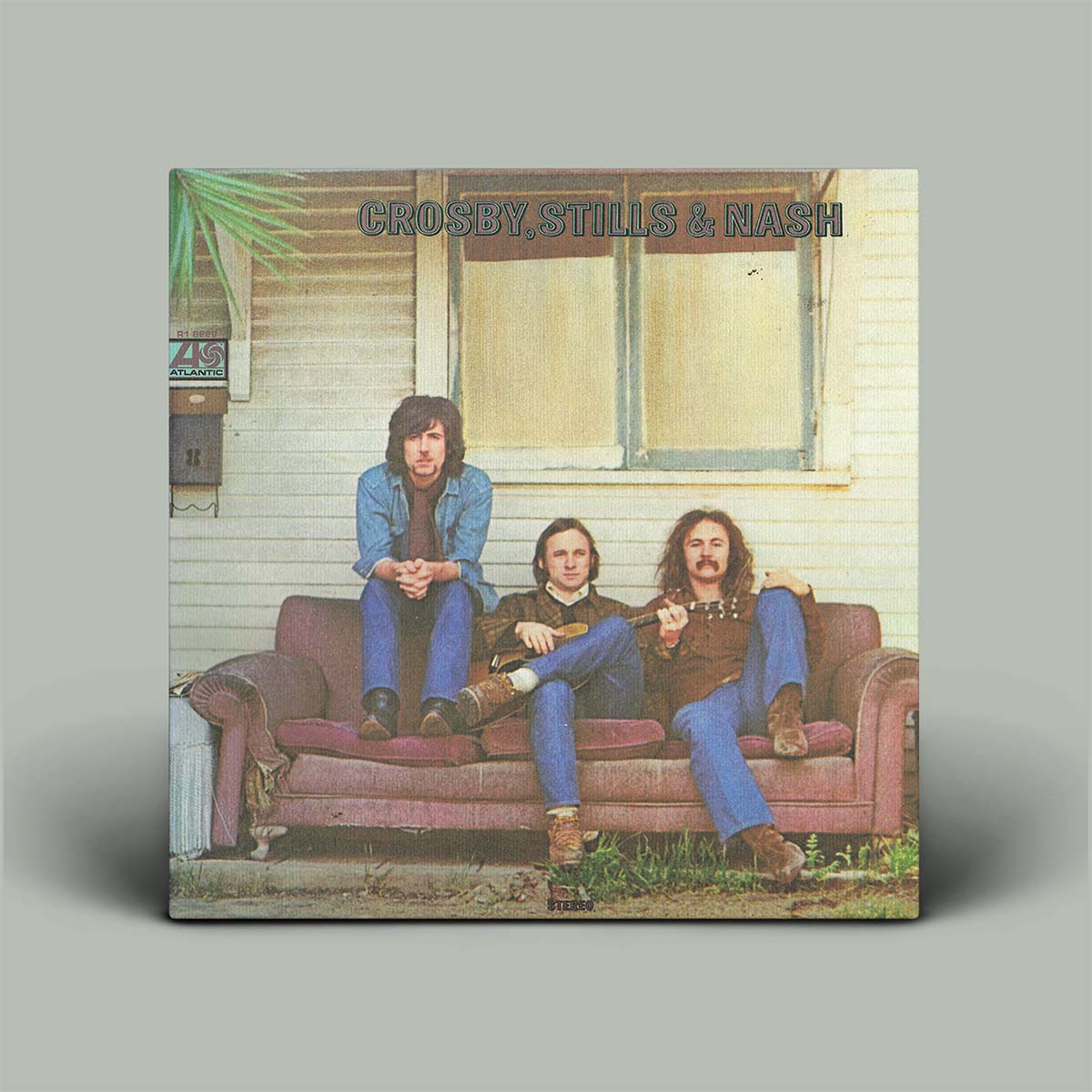 A square album cover for 'Crosby, Stills & Nash' displayed against a solid gray background. The cover features a vintage photograph of the three musicians sitting on a pinkish-brown couch in front of a house with cream siding.