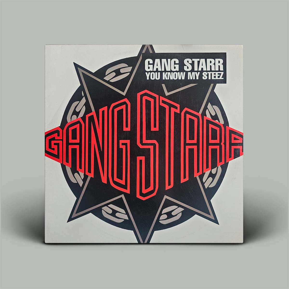 A square album cover for Gang Starr's 'You Know My Steez' is shown standing upright against a plain light gray background. The cover features a large black six-pointed star with red outlined 'GANG STARR' lettering across the center, a black top section with white text, and gray chain graphics.