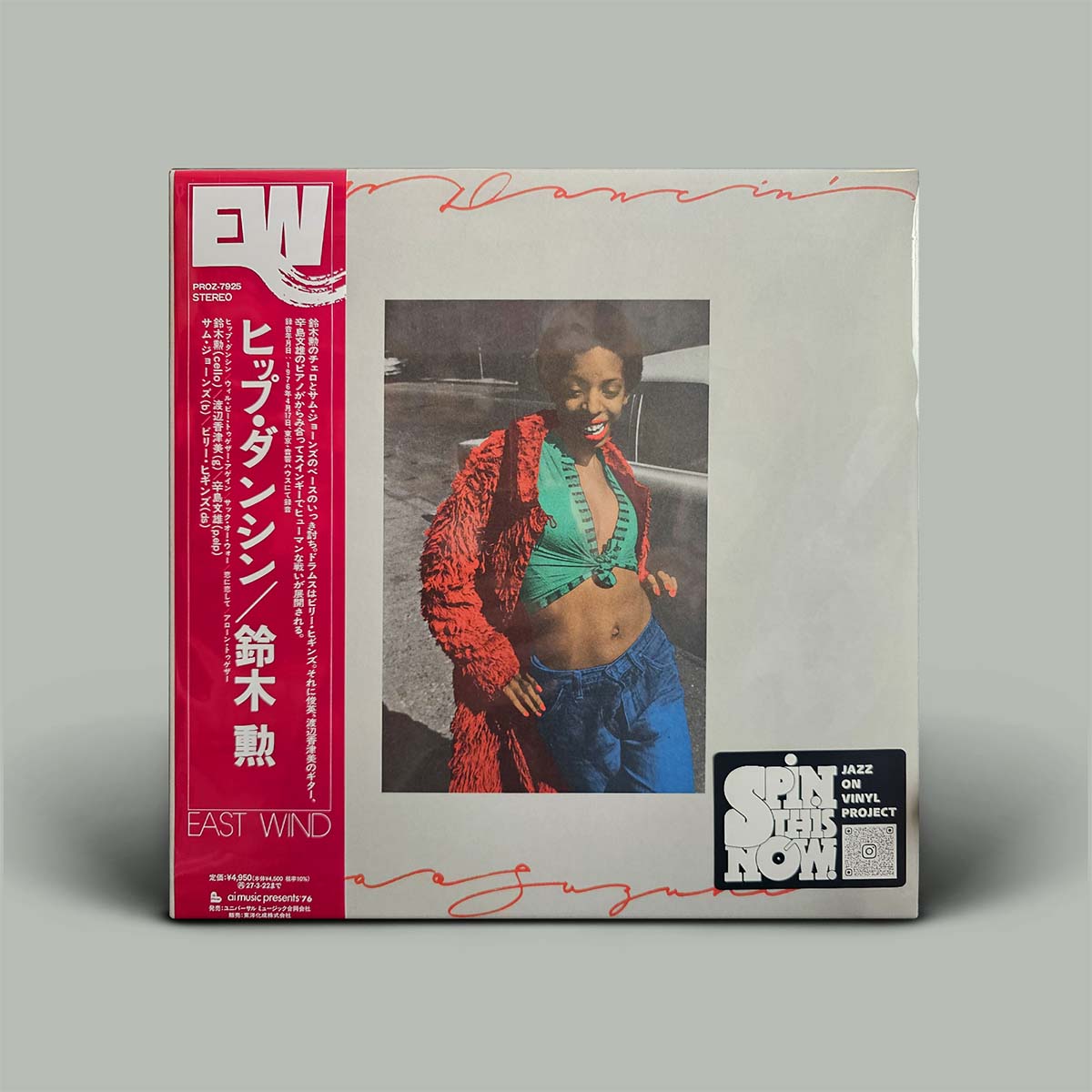 A vinyl record sleeve for an East Wind album featuring a red spine, a central photo of a woman in a red jacket and green top, and a 'Spin It Now' sticker on a gray background.