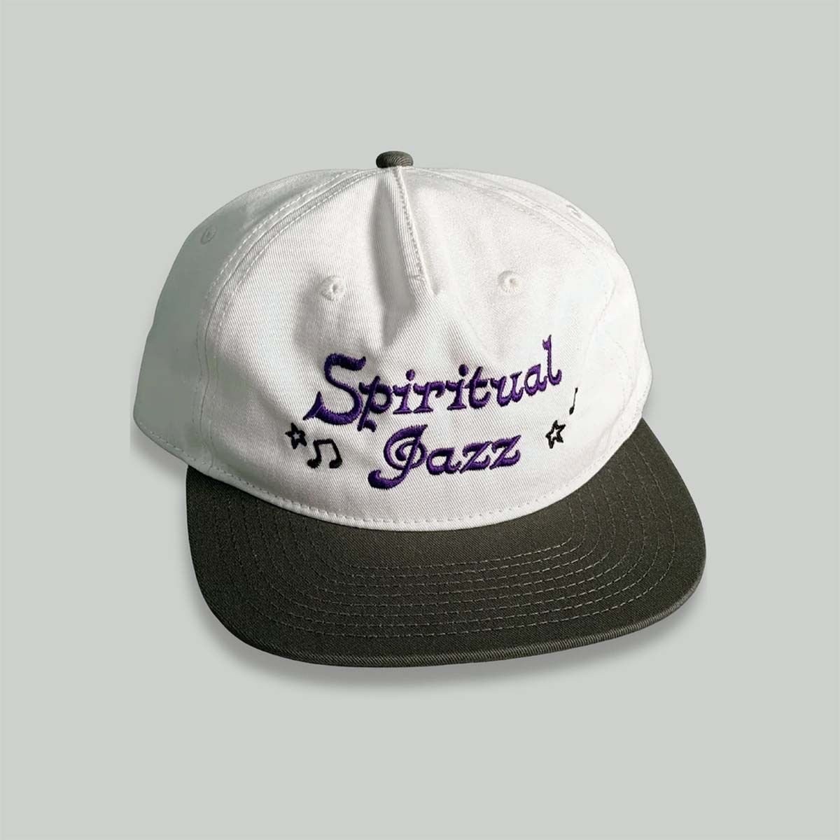 A white snapback cap with a black flat brim, featuring 'Spiritual Jazz' embroidered in purple script on the front, set against a solid light gray background.