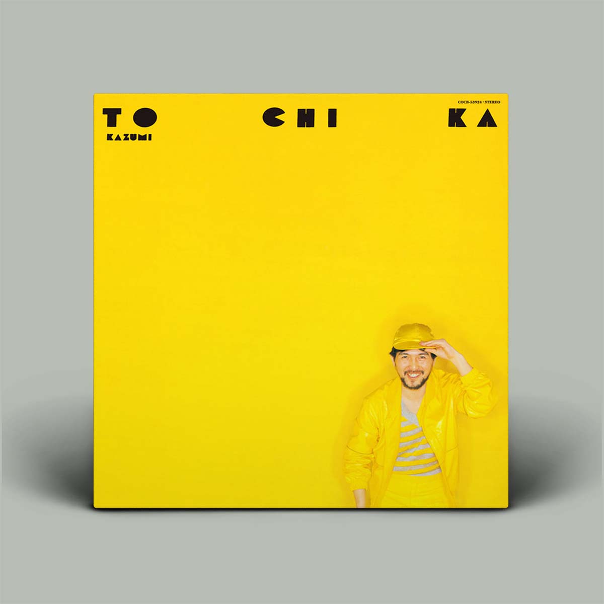 A vinyl record sleeve for album 'TO CHI KA' featuring a solid bright yellow cover with black text and a small portrait of the artist in a yellow outfit at the bottom right, set against a neutral gray background.