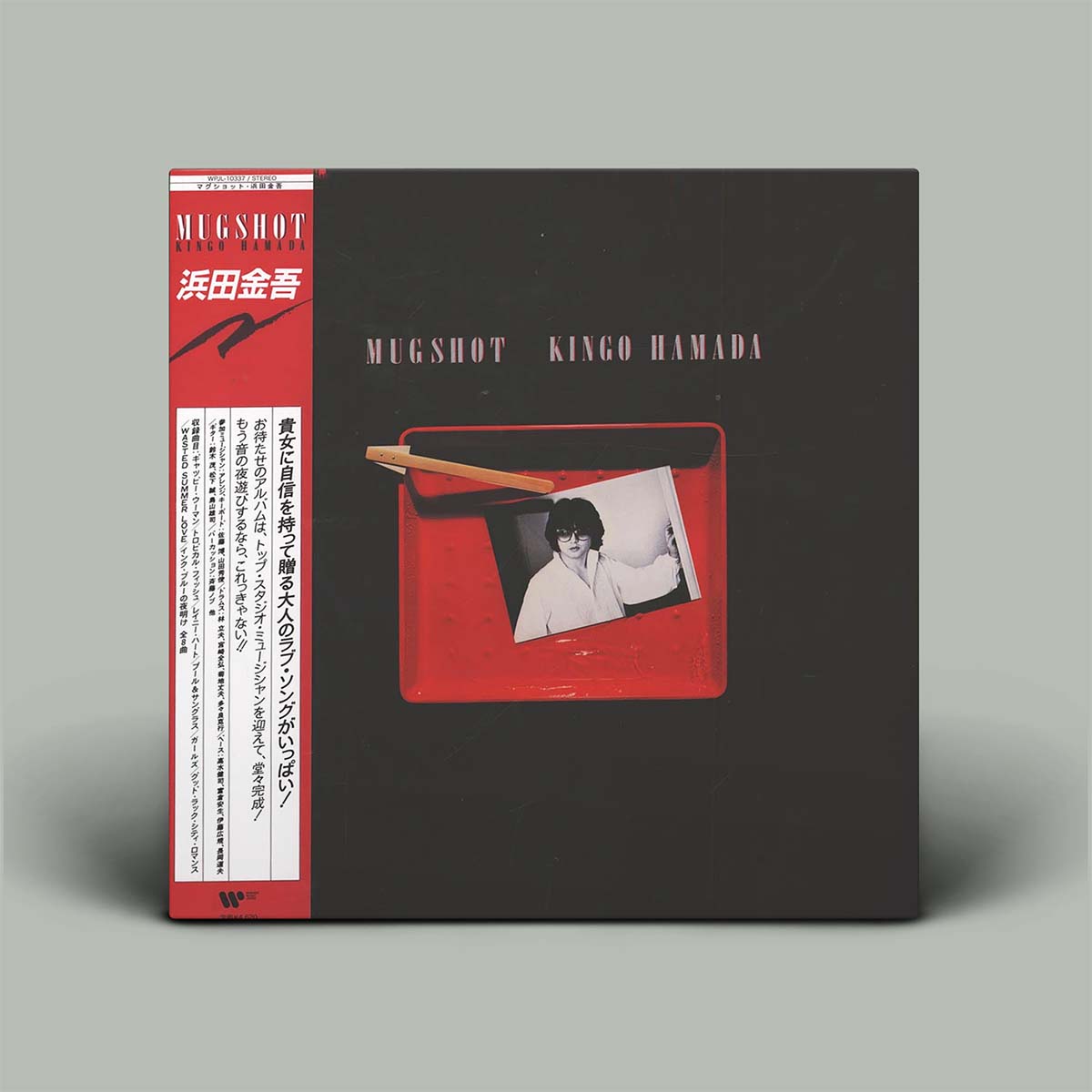 Mugshot Kingo Hamada album cover in a dark charcoal gray color with a red side label and a central red window showing a black-and-white portrait, photographed on a light gray background.