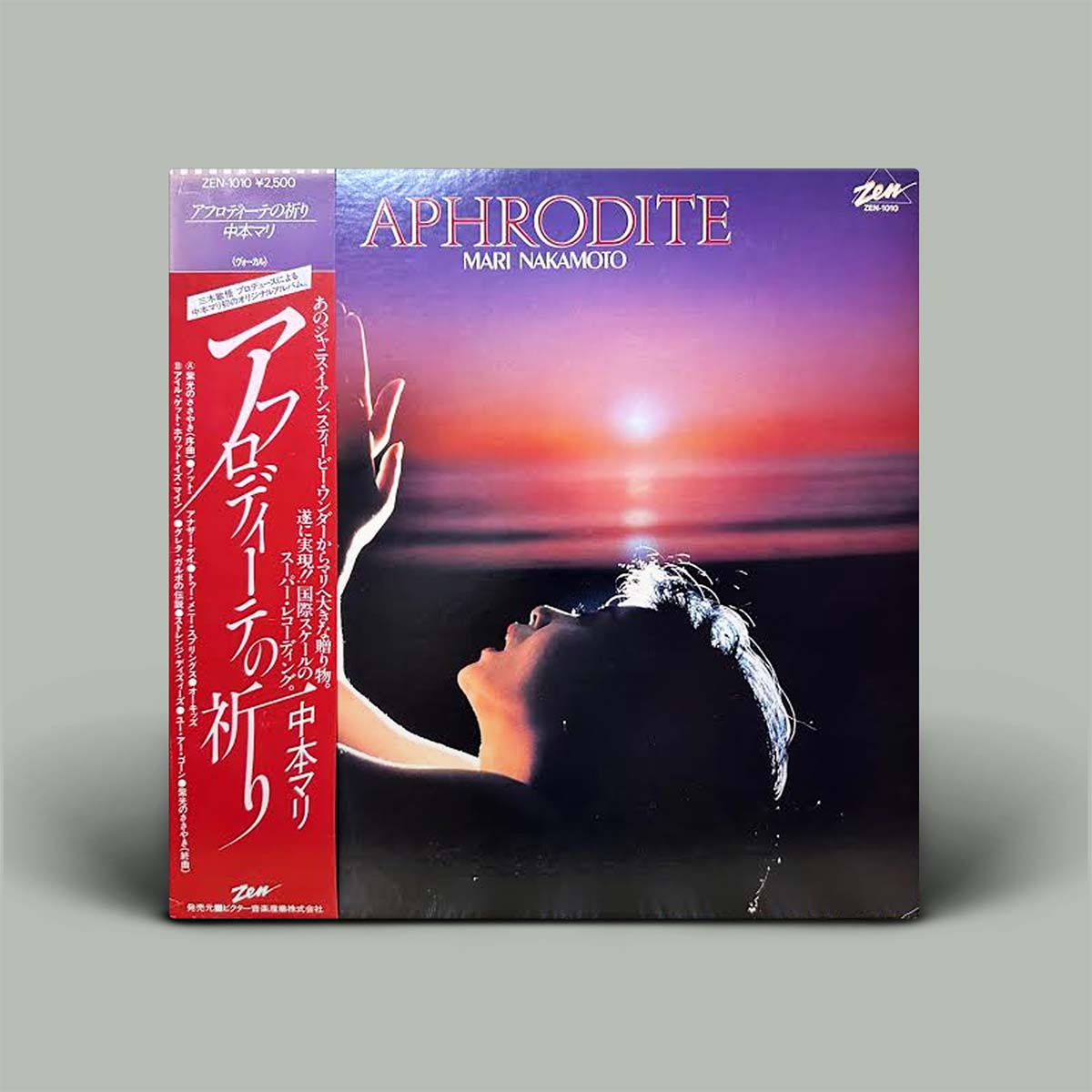 Aphrodite by Mari Nakamori album cover with a red obi strip on the left, featuring a sunset seascape and a woman's profile on the cover, photographed on a gray background.