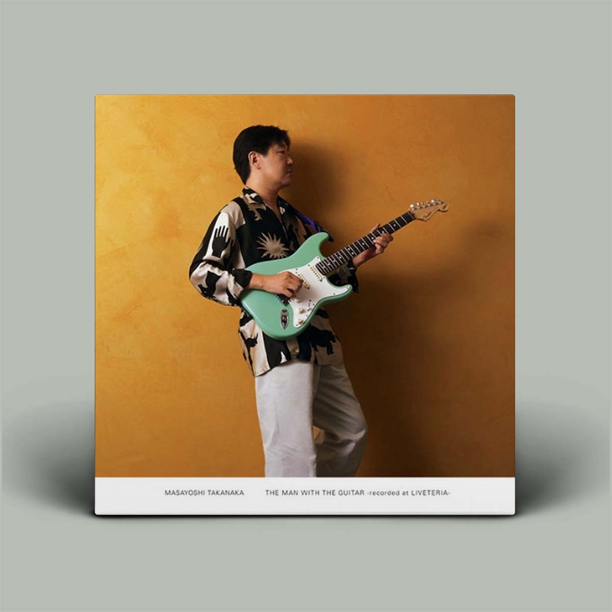 A square album cover for Masayoshi Takanaka's 'The Man With The Guitar' displayed against a neutral gray background. The cover art features a man in a black and white patterned shirt playing a mint green electric guitar against a textured mustard-yellow background, with a white bottom banner containing the album title and artist name.