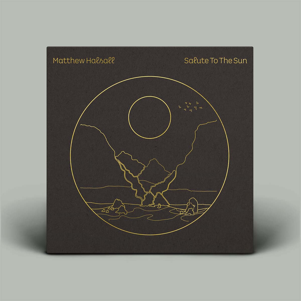 A square dark brown or charcoal album cover standing upright on a light gray background. The cover features gold line art of a minimalist landscape with mountains, water, and a sun, with the text 'Matthew Halebell' and 'Salute To The Sun' in gold at the top.