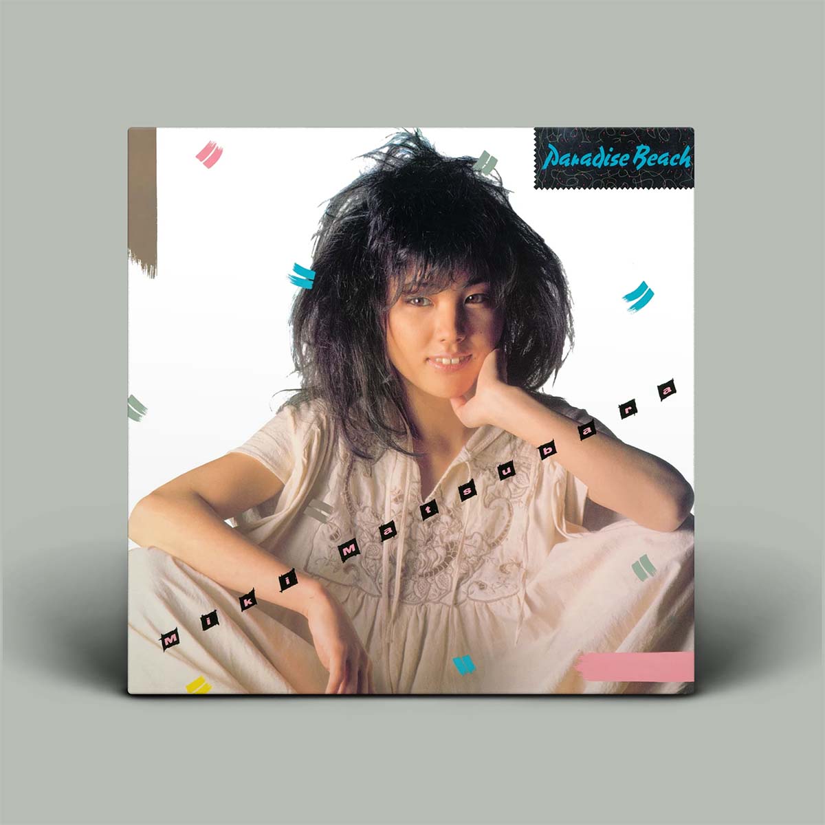 A square album cover for 'Paradise Beach' featuring a woman with dark, voluminous hair in a light beige embroidered dress, set against a white background with colorful geometric accents. The album is shown as a physical square print on a neutral gray background.