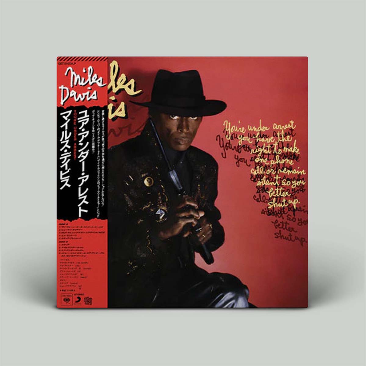 Album cover featuring Miles Davis with a gun in his hand, wearing a black hat and a black coat, against a red background. The text includes the album title 'You're Under Arrest' and the record label.