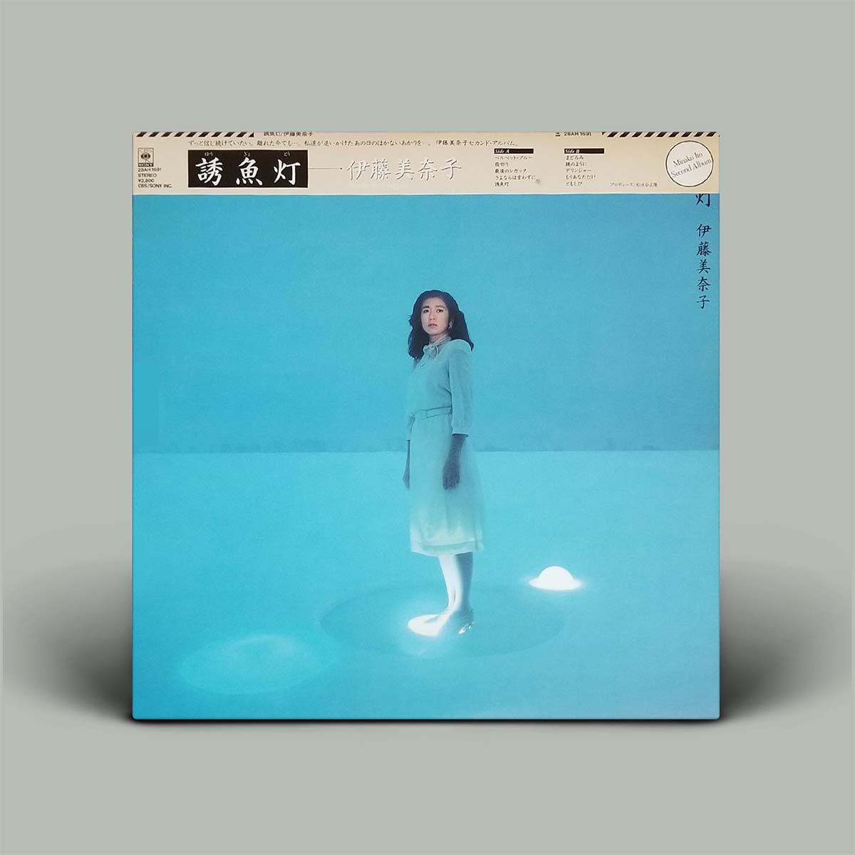 A vinyl record album cover for the artist Minako Ito's Japanese release 'Fish', featuring a photograph of Shimizu standing in a bright blue studio setting with a glowing light beneath her feet. The album is shown standing upright on a neutral gray background.