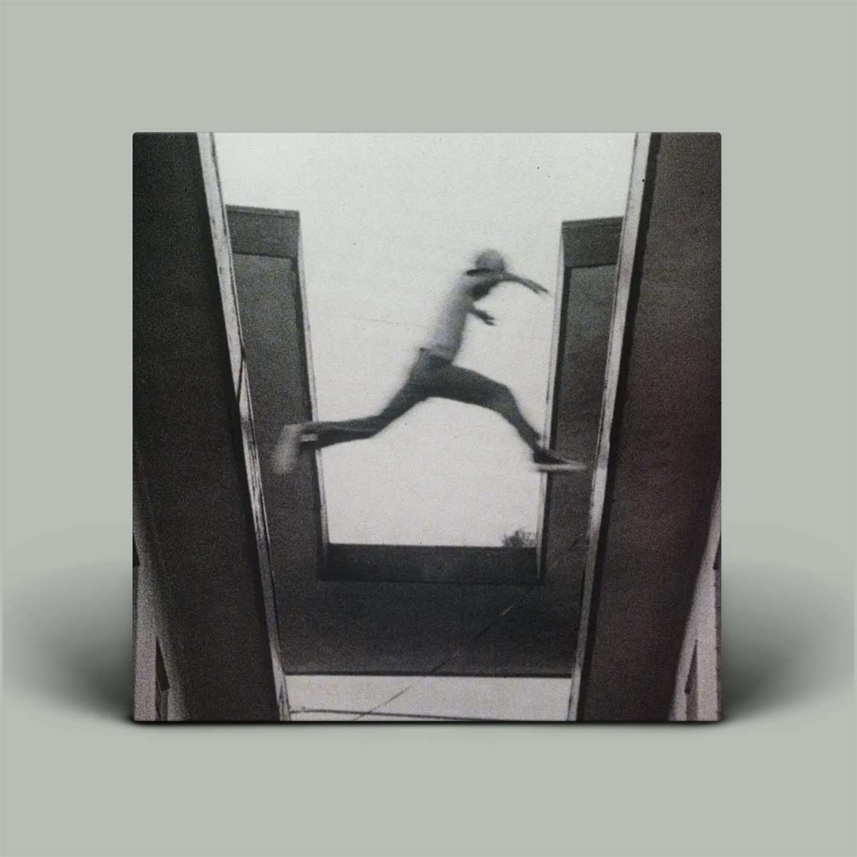 A square black and white photographic art print showing a person mid-jump between two dark vertical structures, displayed as a canvas or framed print against a solid gray background.