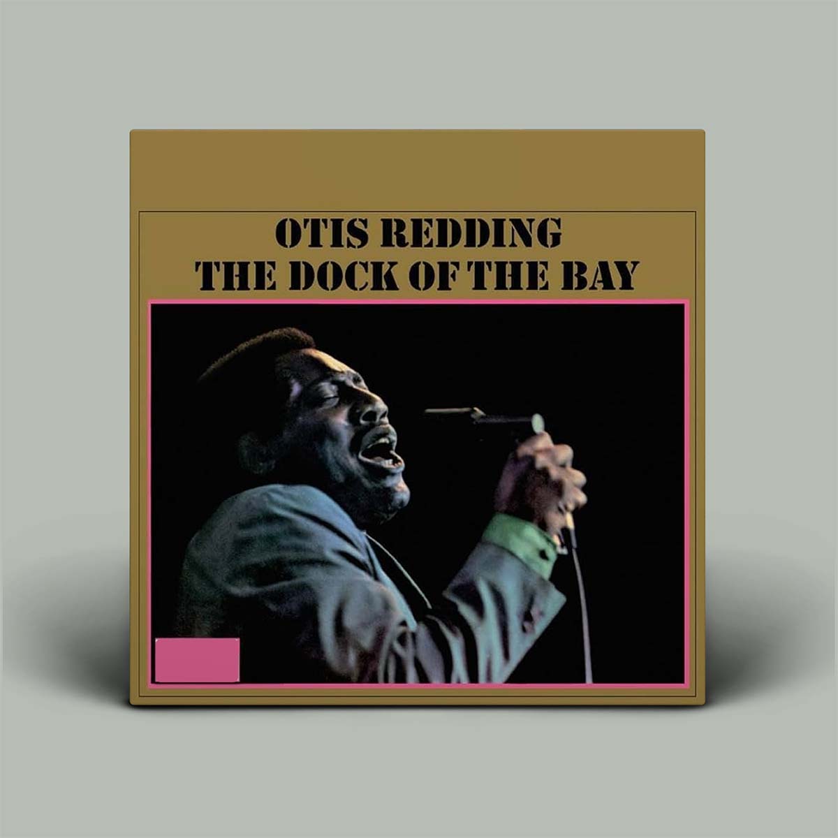 A square album cover for Otis Redding's 'The Dock of the Bay' displayed against a neutral gray background. The cover features a tan border with black text and a central black square containing a photo of Otis Redding singing into a microphone.