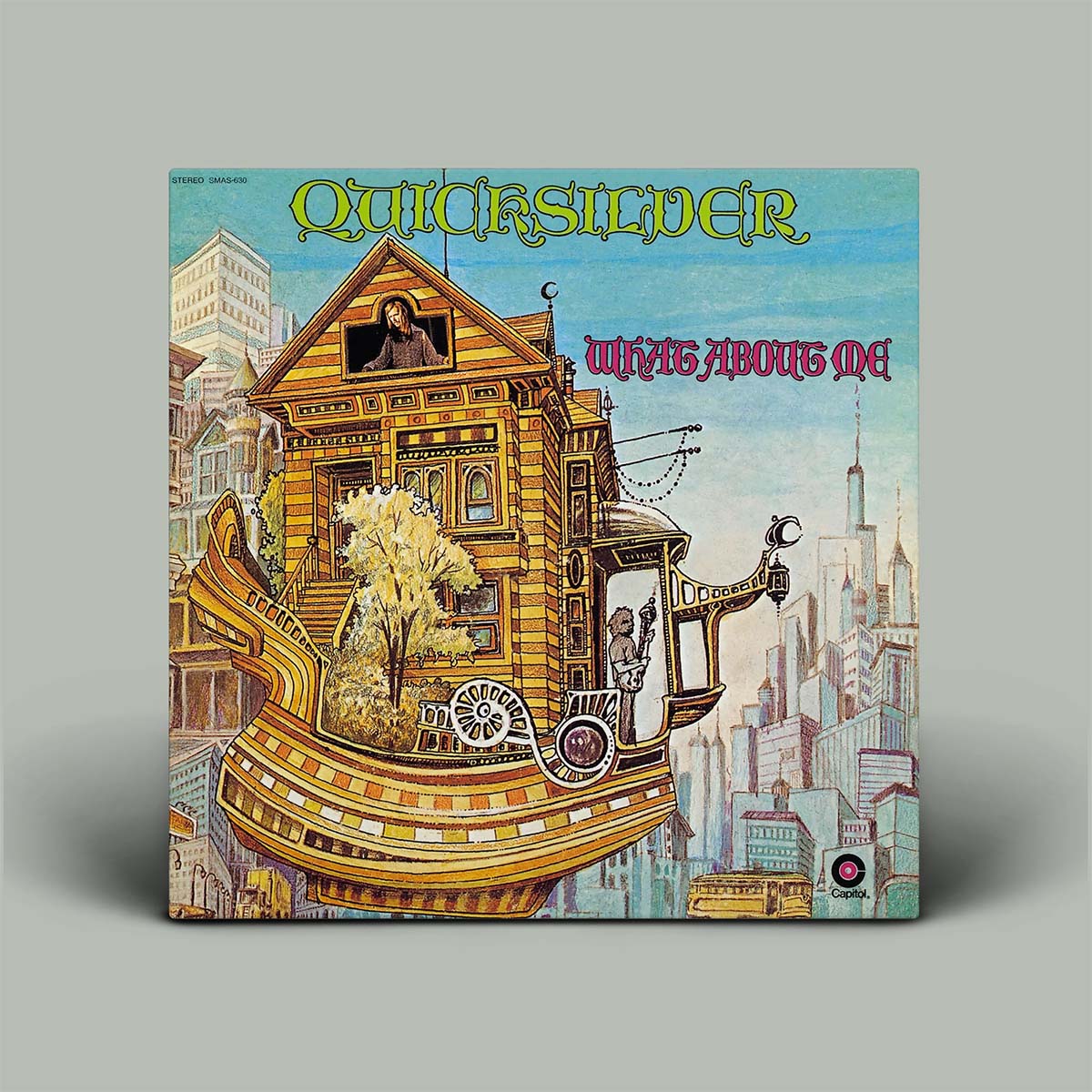 The album cover for Quicksilver's 'WhAT About Me' is shown as a square vinyl record sleeve against a neutral gray background. The artwork features a detailed, whimsical illustration of a house built into a ship's hull, with a small tree in the center, set against a cityscape under a light blue sky.
