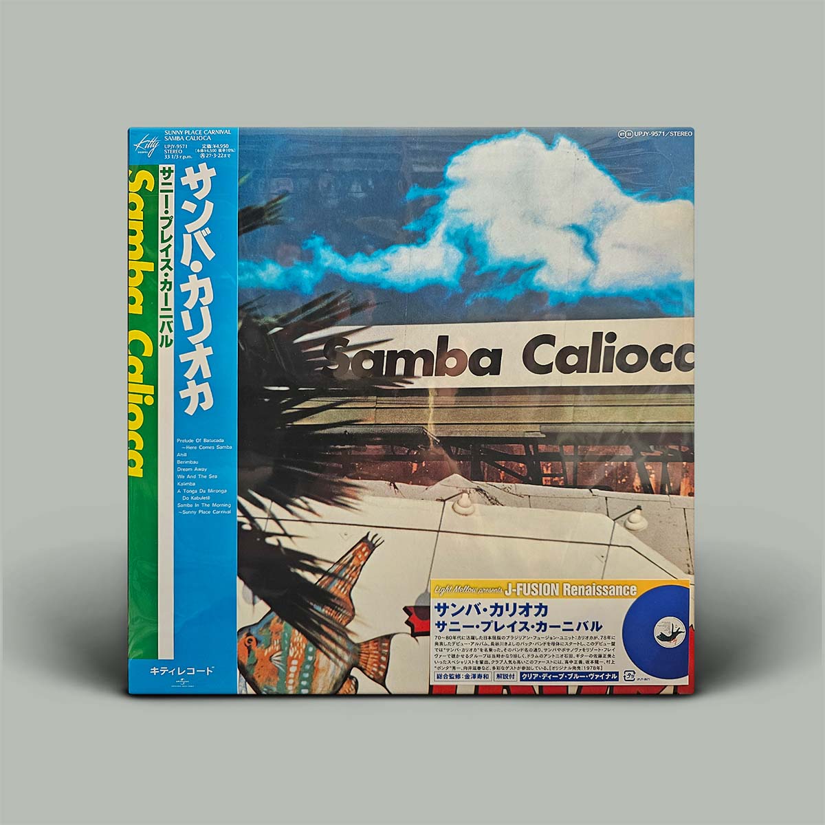 Album cover featuring a tropical beach scene with palm trees, blue sky, and white clouds, shown with its Japanese obi strip on a neutral gray background.