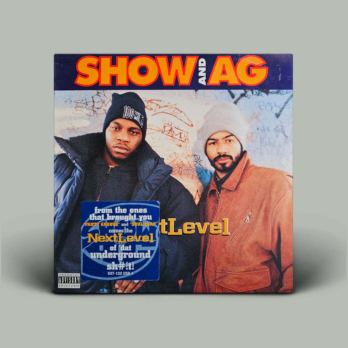 A square vinyl record sleeve for the music release 'Next Level' is shown standing against a solid gray background. The cover art features two men, one in a black puffer jacket and beanie, and the other in a brown jacket and white beanie, with bold orange and white text at the top and a blue sticker overlay on the bottom left.