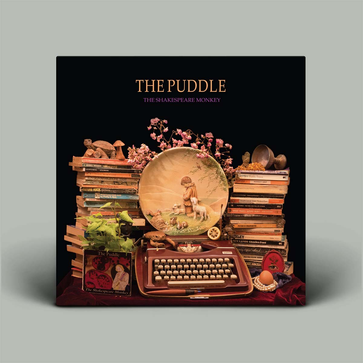 The Puddle - The Shakespeare Monkey album cover, featuring a dark, whimsical still life arrangement with stacks of books, a typewriter, a decorative plate, and a rabbit figurine on a black background.